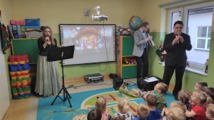 Kobieta w długiej sukni śpiewa do mikrofonu, dwaj muzycy grają na waltorni i trąbce. W tle ekran z wyświetloną prezentacją o świętach Bożego Narodzenia