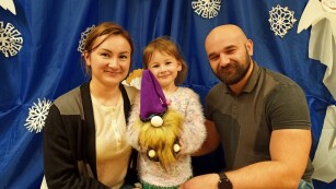Uśmiechnięta dziewczynka z mamą i tatą prezentują wykonanego przez siebie skrzata. W tle świąteczna dekoracja.