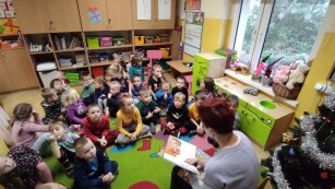 Dzieci siedzą na dywanie, słuchają opowiadania czytanego przez bibliotekarkę