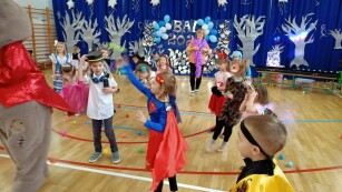 Bal karnawałowy. Sala gimnastyczna udekorowana bibułą, balonami i srebrnymi ozdobami. Dzieci przebrane w stroje karnawałowe uczestniczą w zabawie tanecznej.