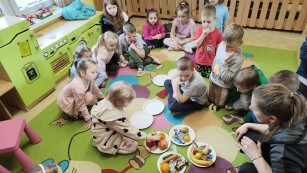 Dzieci siedzą na dywanie. Pośrodku stoją talerzyki, na które nauczycielka wykłada zdrowe i niezdrowe przekąski