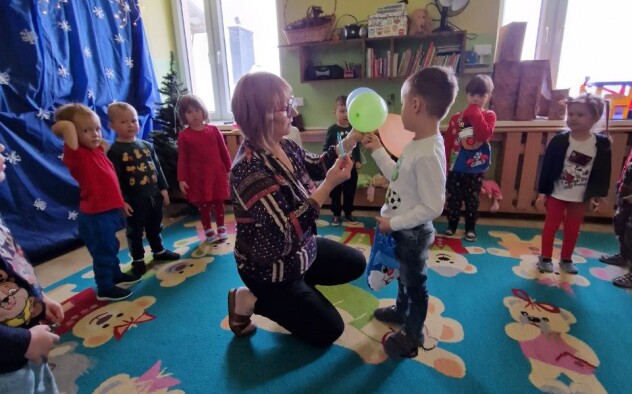 dzieci stoją w kole na sali przedszkolnej. pośrodku nauczycielka wręcza chłopcu balony