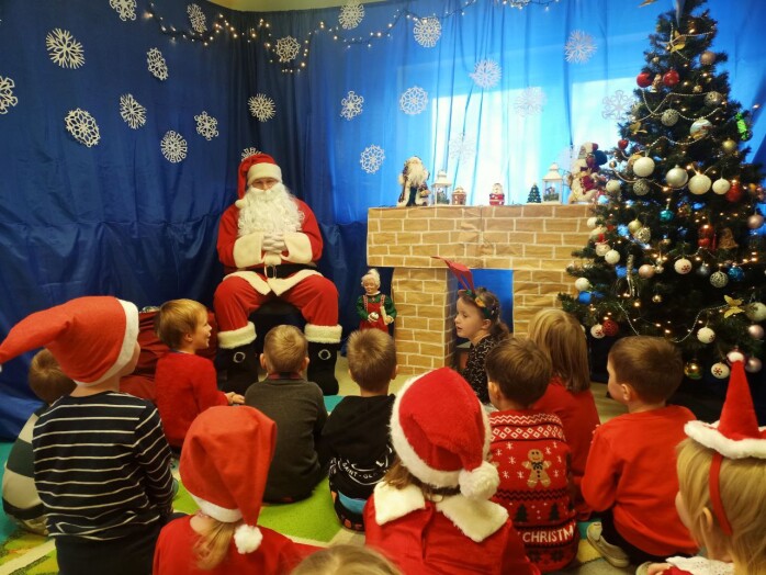 Mikołaj siedzi przy kominku. Przed nim na dywanie siedzą dzieci. W tle granatowe niebo z płatkami śniegu