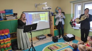 Kobieta śpiewa do mikrofonu, dwaj muzycy grają na waltorni i trąbce pastorałkę Hejże ino