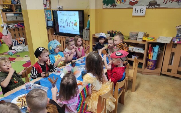 Sala przedszkolna. Dzieci przebrane w stroje karnawałowe uczestniczą siedzą przy stole zastawionym słodkim poczęstunkiem.