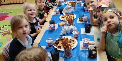 Sala przedszkolna. Dzieci przebrane w stroje karnawałowe uczestniczą siedzą przy stole zastawionym słodkim poczęstunkiem.