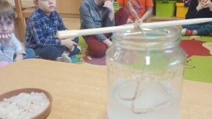 Dzieci siedzą w kole po wykonanym eksperymencie. Na pierwszym planie słoik z roztworem solnym z zanurzonym w nim sznurkiem.