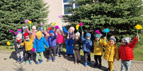 Zdjęcie grupowe. Dzieci z kolorowymi kwiatami z bibuły stoją przed budynkiem przedszkola
