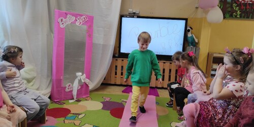 Dziecko prezentuje strój Barbie na dziecięcym pokazie mody. Idzie po różowym dywanie, po bokach siedzi dziecięca publiczność