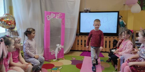 Dziecko prezentuje strój Barbie na dziecięcym pokazie mody. Idzie po różowym dywanie, po bokach siedzi dziecięca publiczność