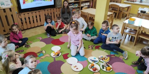 Dzieci siedzą na dywanie. Pośrodku stoją talerzyki, na które dziewczynka wykłada zdrowe i niezdrowe przekąski