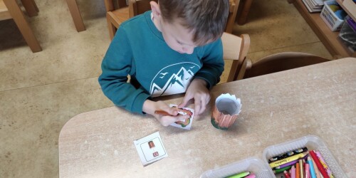 Chłopiec siedzi przy stoliku. Rysuje kredkami etykietę do doniczki z kasztanowcem.